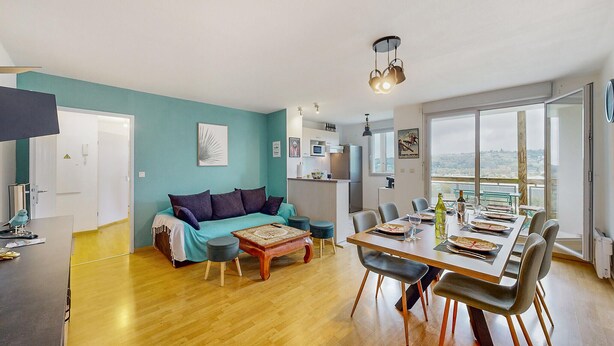 Ein modernes Wohnzimmer mit einem türkisblauen Sofa, einem Holztisch und einem Essbereich mit einem großen Fenster mit Blick auf die Stadt.