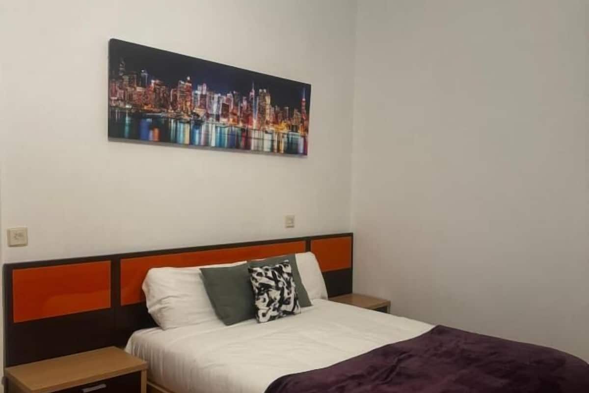 A bedroom with a bed, nightstands, a cityscape painting, and a ceiling light.