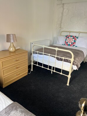 A bedroom with a metal-framed bed, a wooden dresser with drawers, a lamp, and a radiator. - Cozy Gogarstone Cottage in Newbridge (Newbridge)