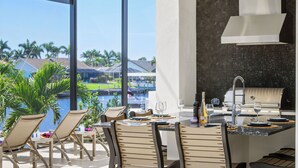 A dining area with a view of a pool and palm trees, a grill, and outdoor seating.