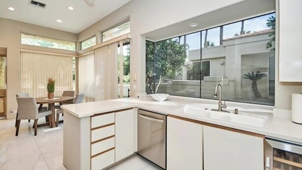 A modern kitchen with a large island, a dining area with a table and chairs, and a view of the outdoors through large windows.