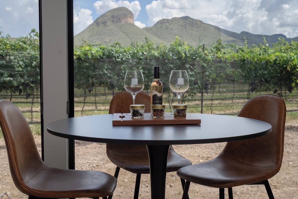 A table with two wine glasses, a bottle, and small jars, set against a vineyard backdrop with mountains.