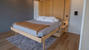 Une chambre moderne avec un cadre de lit en bois, un matelas gris et un plaid marron. On y trouve une table de chevet en bois, une lampe et une prise électrique.