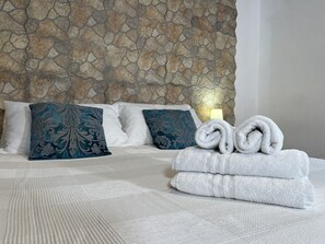 A neatly made bed with white bedding and decorative pillows, a stone wall behind, and a folded towel on the bed.
