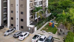 Un edificio de varios pisos con el nombre 'GOSAIN' en la fachada, con balcones, un estacionamiento con varios autos y vistas a zonas verdes y colinas distantes.