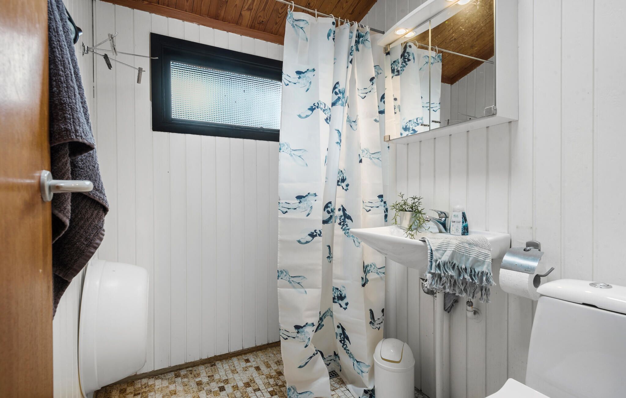 A bathroom with a shower curtain featuring a blue floral pattern, a white sink with a towel, and a window above the toilet.