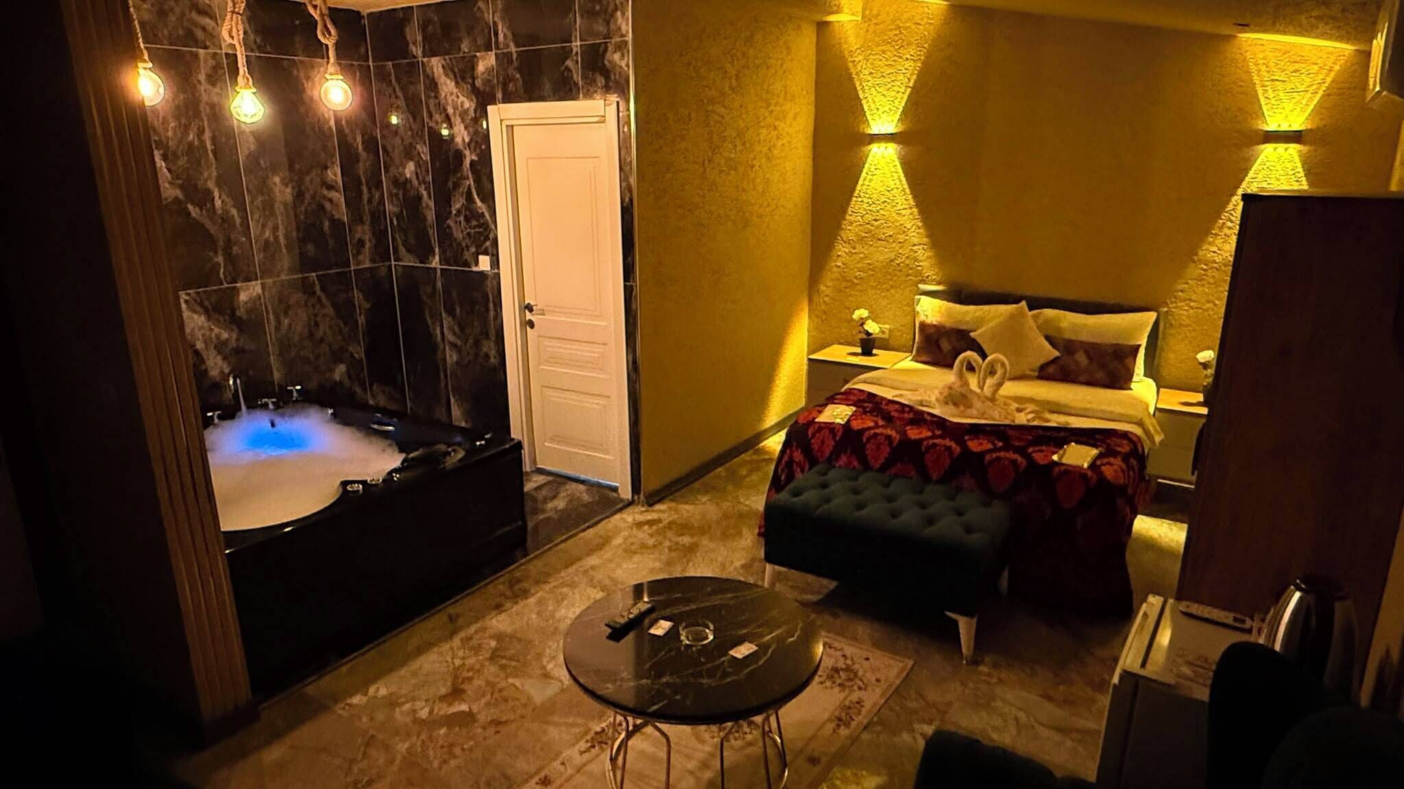 A room with a bathtub, a bed with a tufted headboard, a small round table, and a nightstand with a lamp.