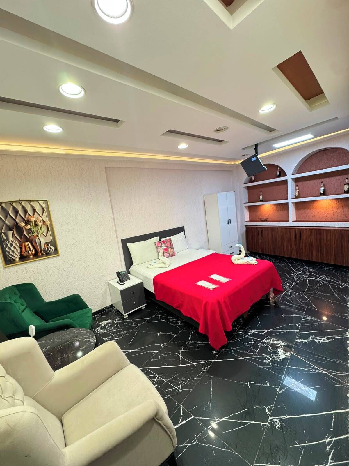 Une chambre d’hôtel avec un lit, un fauteuil vert, un canapé blanc, un minibar et une étagère fixée au mur, sur laquelle sont posées des bouteilles.