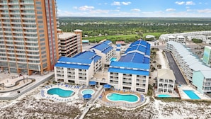 A high-rise building with multiple balconies, a swimming pool area with several pools, and a view of a green landscape in the background. - Cozy Gulf-Side Studio Sugar Beach 235 (Orange Beach)