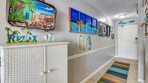 A hallway with a white textured cabinet, a colorful striped rug, and various wall art including a beach scene and a seahorse. - Cozy Gulf-Side Studio Sugar Beach 235 (Orange Beach)
