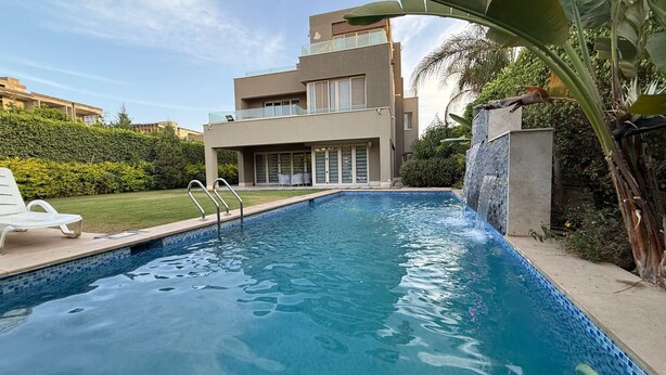 A swimming pool with a modern house in the background, a small waterfall feature, and a well-maintained lawn. - 6-bedroom villa with pool & garden in palm hills-new cairo (Cairo Governorate)