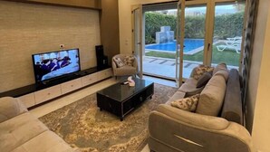 A modern living room with a large TV, comfortable seating, and a view of a swimming pool. - 6-bedroom villa with pool & garden in palm hills-new cairo (Cairo Governorate)