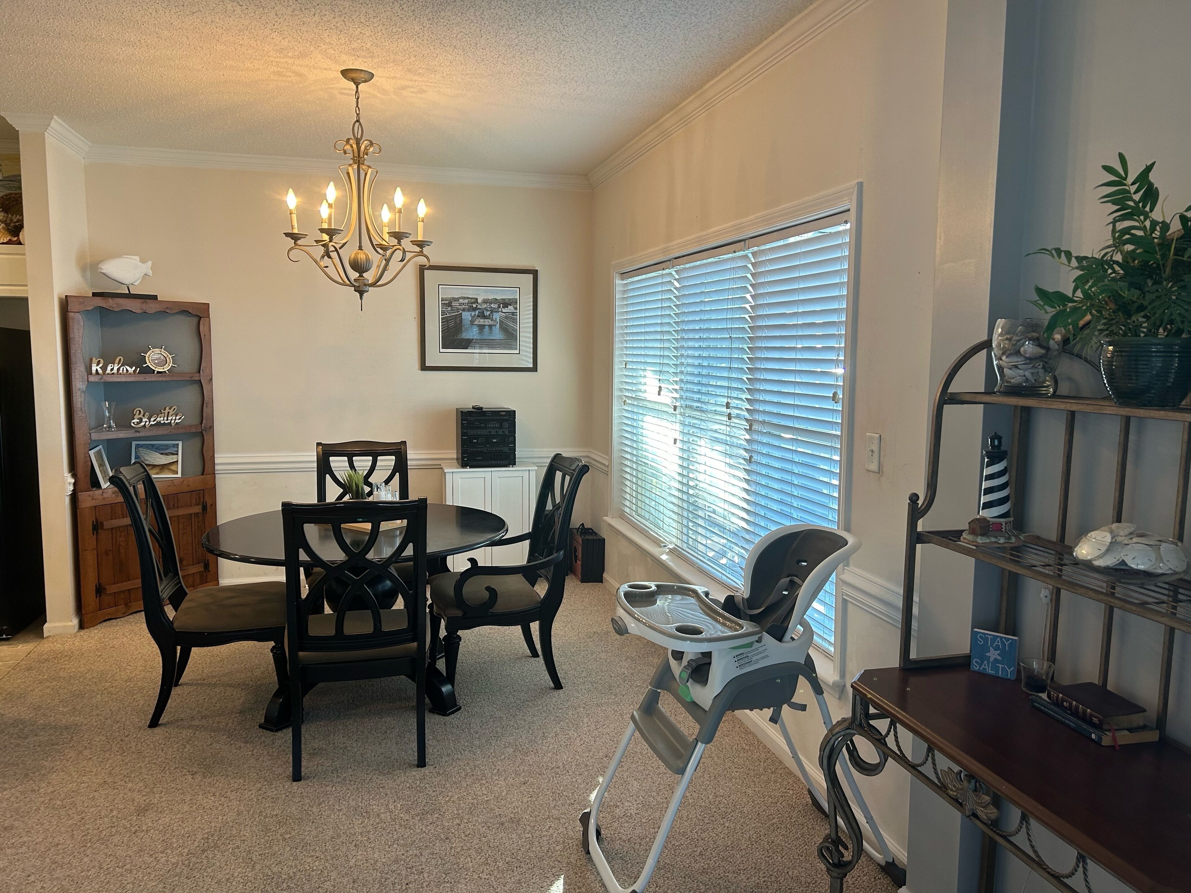 A dining area with a chandelier, a round table, and chairs. There is a window with blinds, a shelf with decorative items, and a sign that says "STAY SALTY".