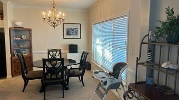 A dining area with a chandelier, a round table, and chairs. There is a window with blinds, a shelf with decorative items, and a sign that says "STAY SALTY".