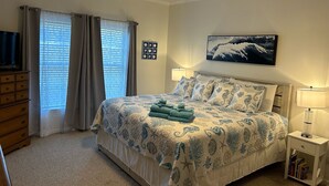A bedroom with a bed, a ceiling fan, a window with curtains, a wall-mounted picture, and a bedside table with a lamp and books.