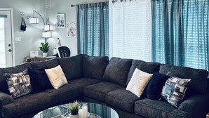 A living room with a dark gray sectional sofa, a glass coffee table with decorative fish, a ceiling fan, and a view of the outdoors through large windows with blue curtains.
