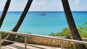 A view of a clear blue sea with a boat in the distance, framed by palm trees and a stone wall. - Beachfront Breeze Condo (Philipsburg)