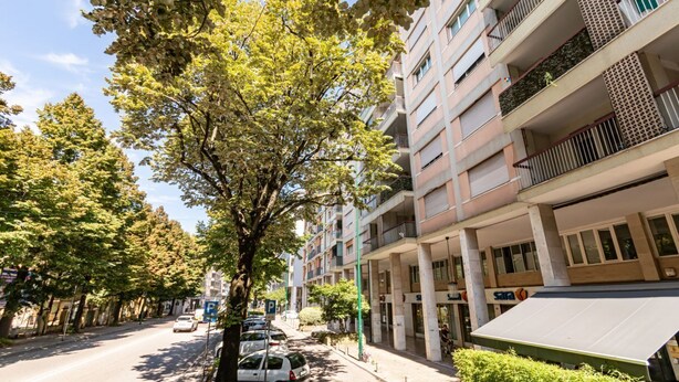 Un edificio residencial de varios pisos con balcones, una zona de estacionamiento y árboles a lo largo de la calle.