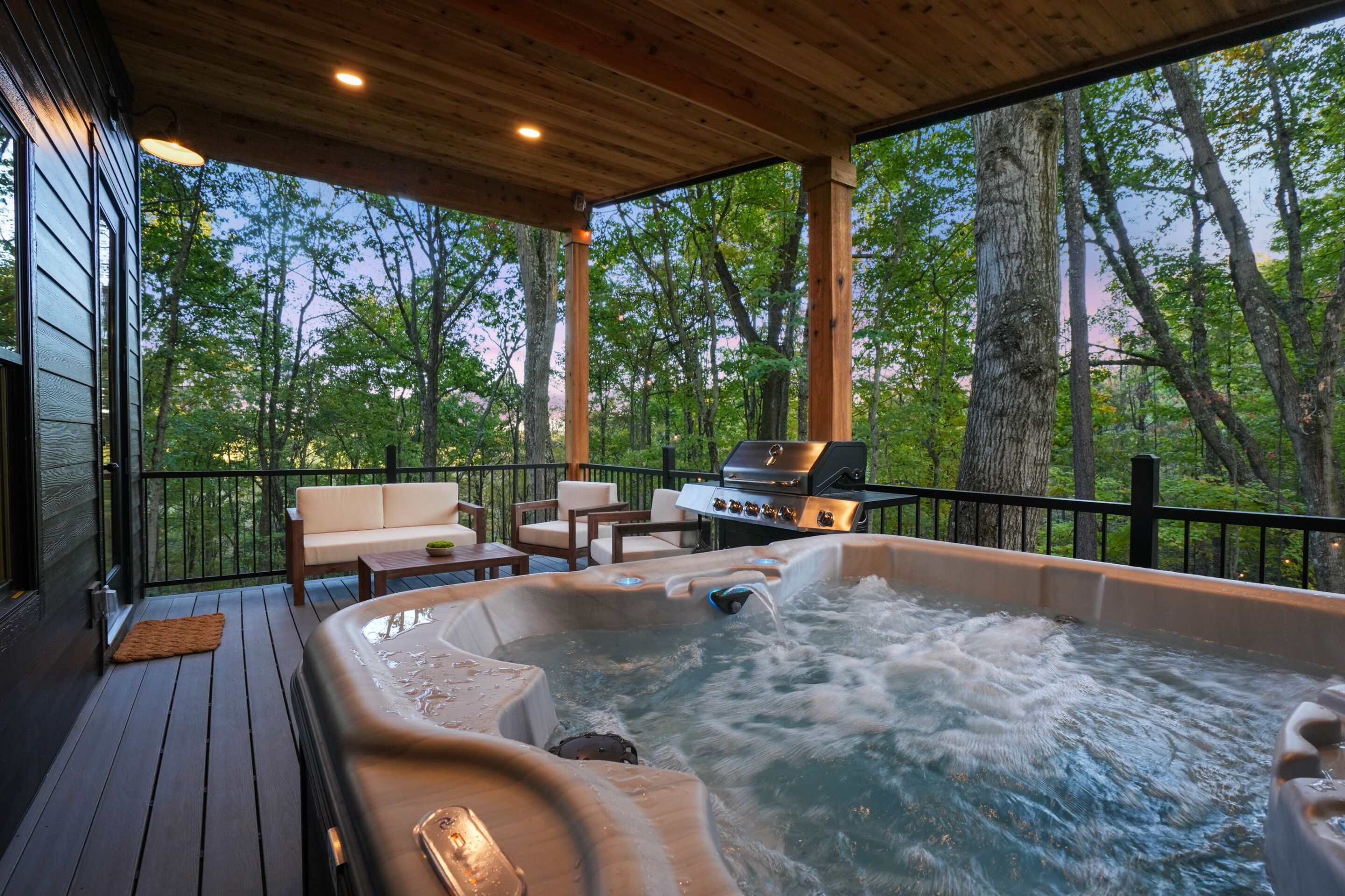 Un bain à remous sur une terrasse en bois, avec un barbecue et un espace détente, le tout entouré d’arbres.