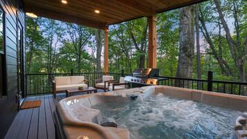 Un bain à remous sur une terrasse en bois, avec un barbecue et un espace détente, le tout entouré d’arbres.