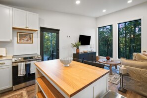 Une cuisine moderne avec des meubles blancs, un îlot central en bois et un four encastré. On y trouve un coin repas avec une table ronde et des chaises, ainsi qu’un salon avec un canapé beige et un téléviseur fixé au mur.