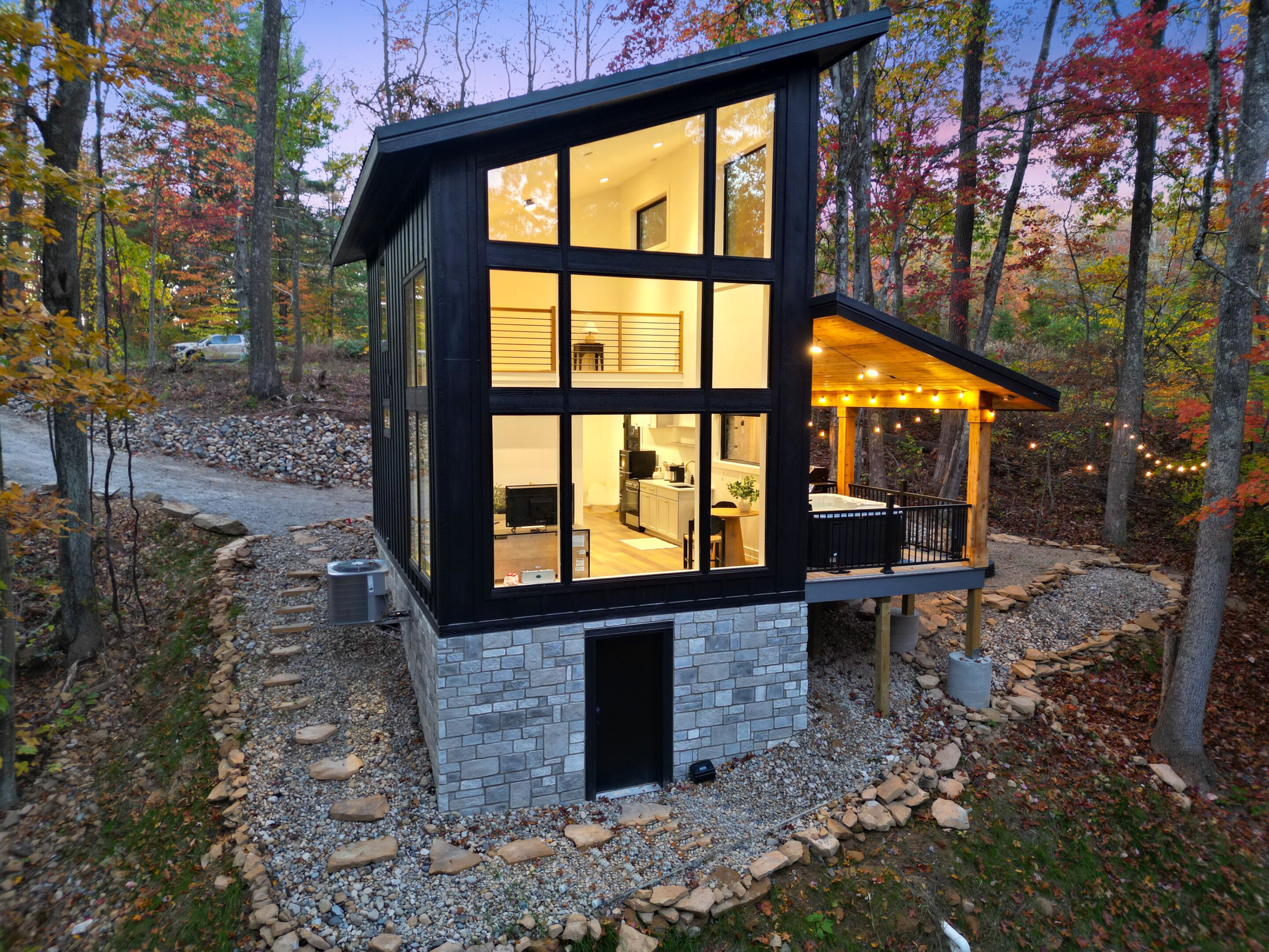 A modern two-story cabin with large windows, a stone foundation, and a deck with string lights.