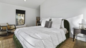 A modern bedroom with a large bed, a chair, a nightstand, and a window with a view of trees.