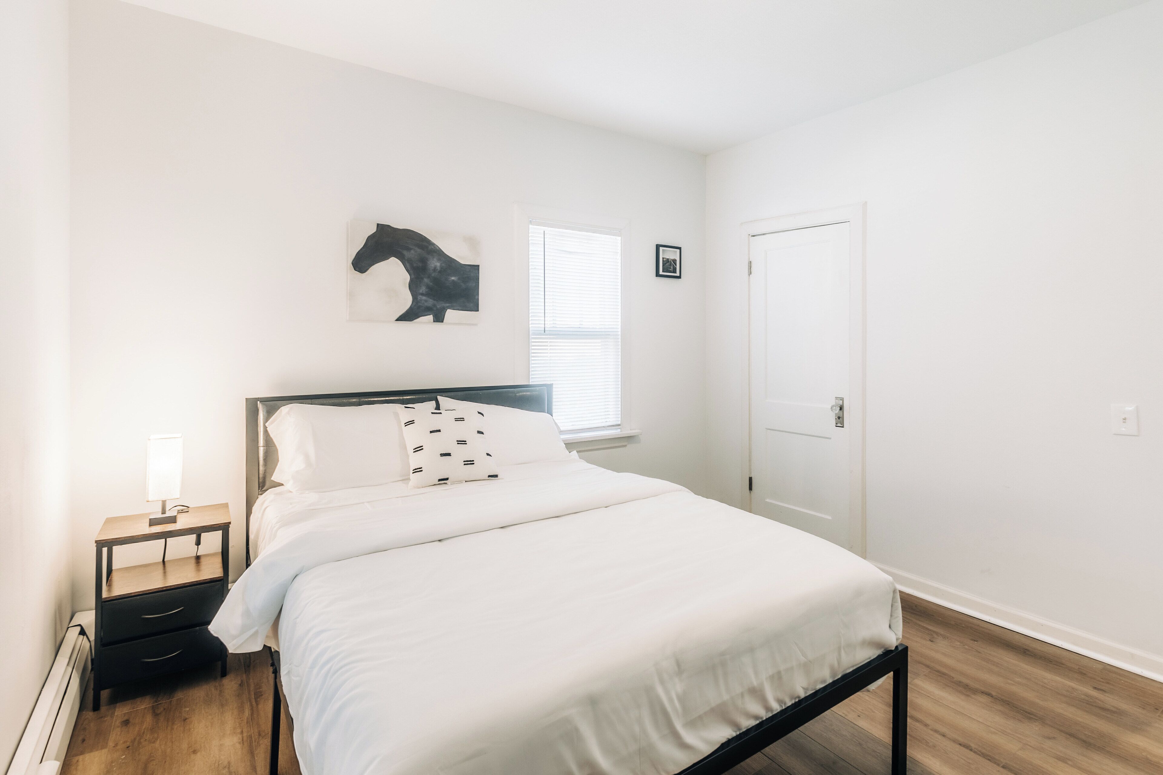 A bedroom with a bed, a nightstand, a wall-mounted artwork, a window, and a door.