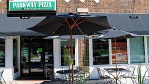 A brick building with a sign for Parkway Pizza. There are outdoor tables and chairs under umbrellas.