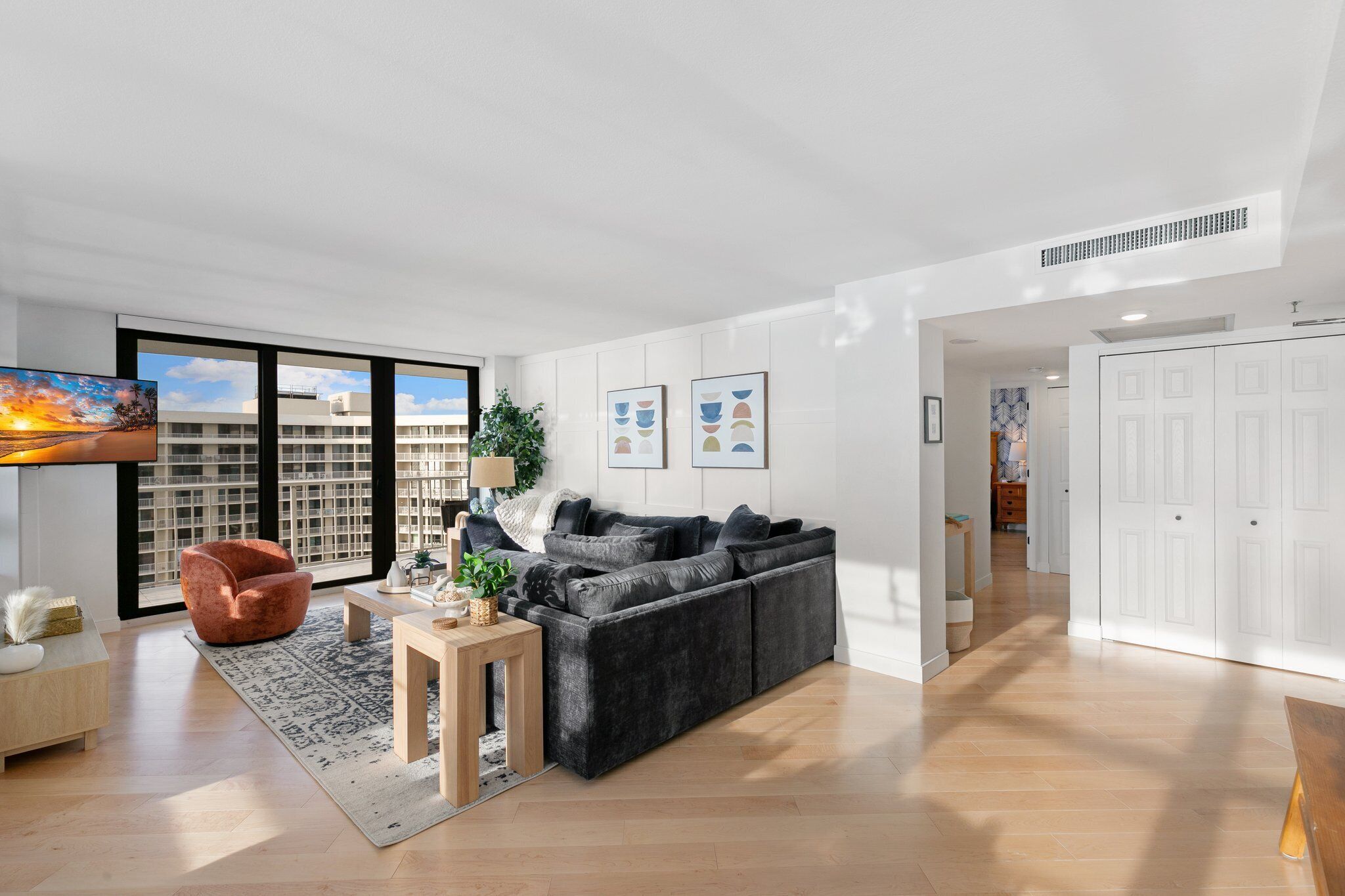 A modern living room with a grey sectional sofa, a wooden coffee table, a red armchair, and a large window with a city view.