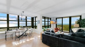 A spacious living area with a large window overlooking a scenic view, a dining area with a table set for two, and a cozy seating area with a sofa. - Stylish Front Corner Beachfront Resort | 5⭐️ Local Host | Balcony | Views (Marco Island)