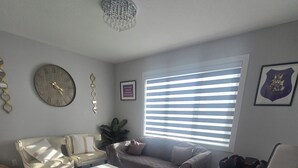 A living room with a grey sofa, a white coffee table, a black armchair, a chandelier, a clock, and a window with blinds.