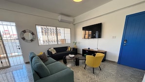 Un salon avec un canapé, une table basse et un fauteuil jaune. Un téléviseur est fixé au mur et il y a une porte bleue.