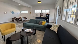 Un salon spacieux comprenant un canapé, un fauteuil et une table basse. On y trouve également un coin repas avec des chaises et une table. La pièce est dotée de grandes fenêtres et d'un luminaire au plafond.