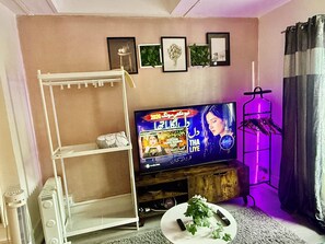 Una sala de estar con televisor, estantería blanca, planta y perchero.