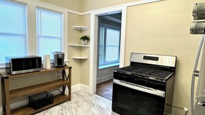 A kitchen with a microwave, coffee maker, and stove. There are windows with blinds, a shelf with a plant, and a wooden table with a basket.