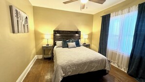 A bedroom with a wooden headboard, two bedside tables with lamps, a bed with a patterned bedspread, and a window with curtains.