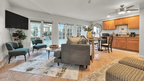 Un vaste espace de vie comprenant un canapé gris, deux fauteuils, une table basse et une table à manger avec des chaises. Il y a également un coin cuisine avec des meubles en bois et un ventilateur de plafond.