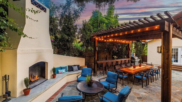 Une terrasse avec un sol en pierre, une cheminée, des sièges rembourrés bleus et une pergola illuminée de guirlandes lumineuses.
