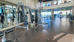 Un gimnasio amplio con diversos equipos de ejercicio, un entrepiso y grandes ventanales.