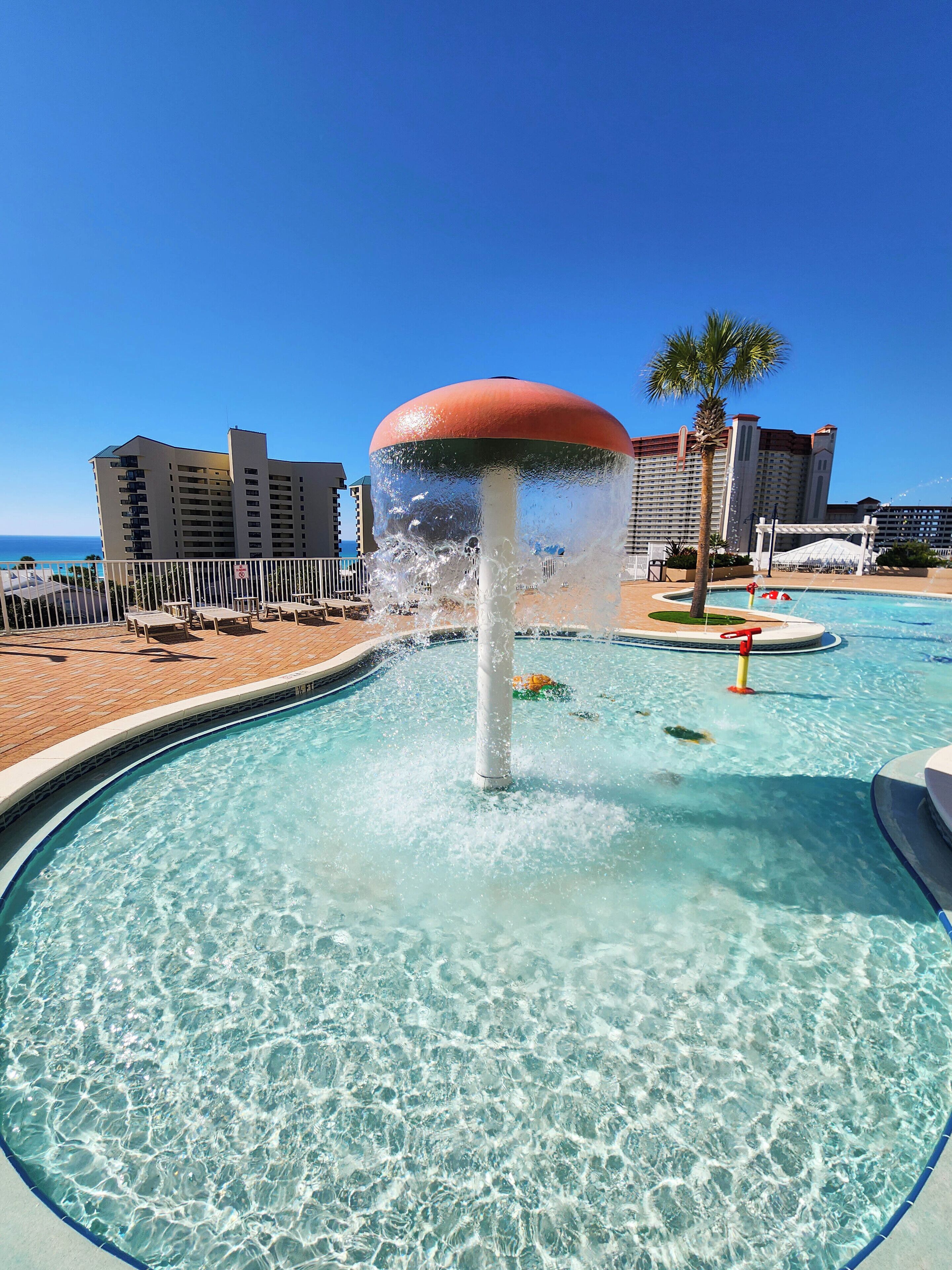 Ein Pool mit einer Springbrunnenanlage, umgeben von einer Poolterrasse mit Liegestühlen und einer Palme.