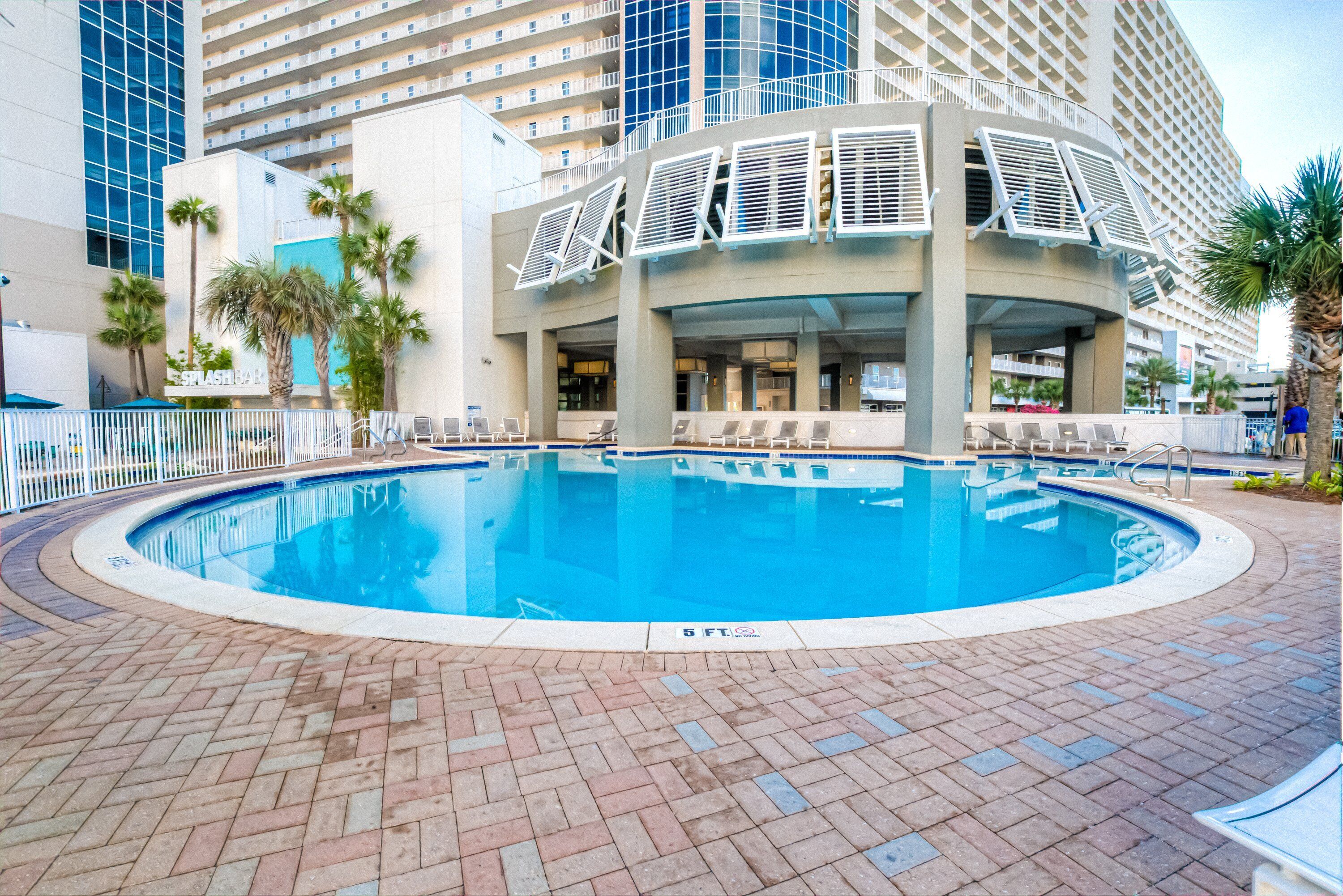 Piscina con borde de ladrillo, rodeada de sillas de descanso y palmeras, con un edificio de hotel al fondo.
