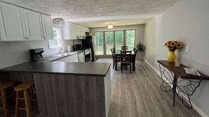 A modern kitchen with a central island, bar stools, and a dining area with a table and chairs. - Cozy Modern Cabin in the Woods (Hawley)