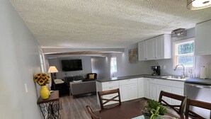 A modern kitchen with white cabinets, a dining table with chairs, and a small side table with a lamp and flowers. - Cozy Modern Cabin in the Woods (Hawley)