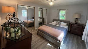 A bedroom with a bed, a nightstand, a lamp, a mirror, and a window. - Cozy Modern Cabin in the Woods (Hawley)