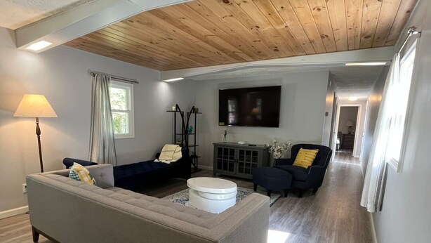 A spacious living room with a high ceiling, wooden beams, a grey sofa, a dark armchair, a white coffee table, and a flat-screen TV mounted on the wall. - Cozy Modern Cabin in the Woods (Hawley)