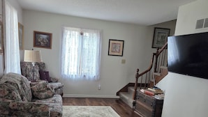 A living room with a floral sofa, a wooden coffee table, a patterned rug, a staircase, and a flat-screen TV mounted on the wall.
