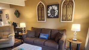 A cozy living room with a brown sofa, a grey armchair, and a blue 'Welcome' pillow. There are two wall-mounted lamps and a small table with a lamp. - 4BR Connected Condo Near Silver Dollar City | Great for Groups | NCH 7 7-8 (Reeds Spring)