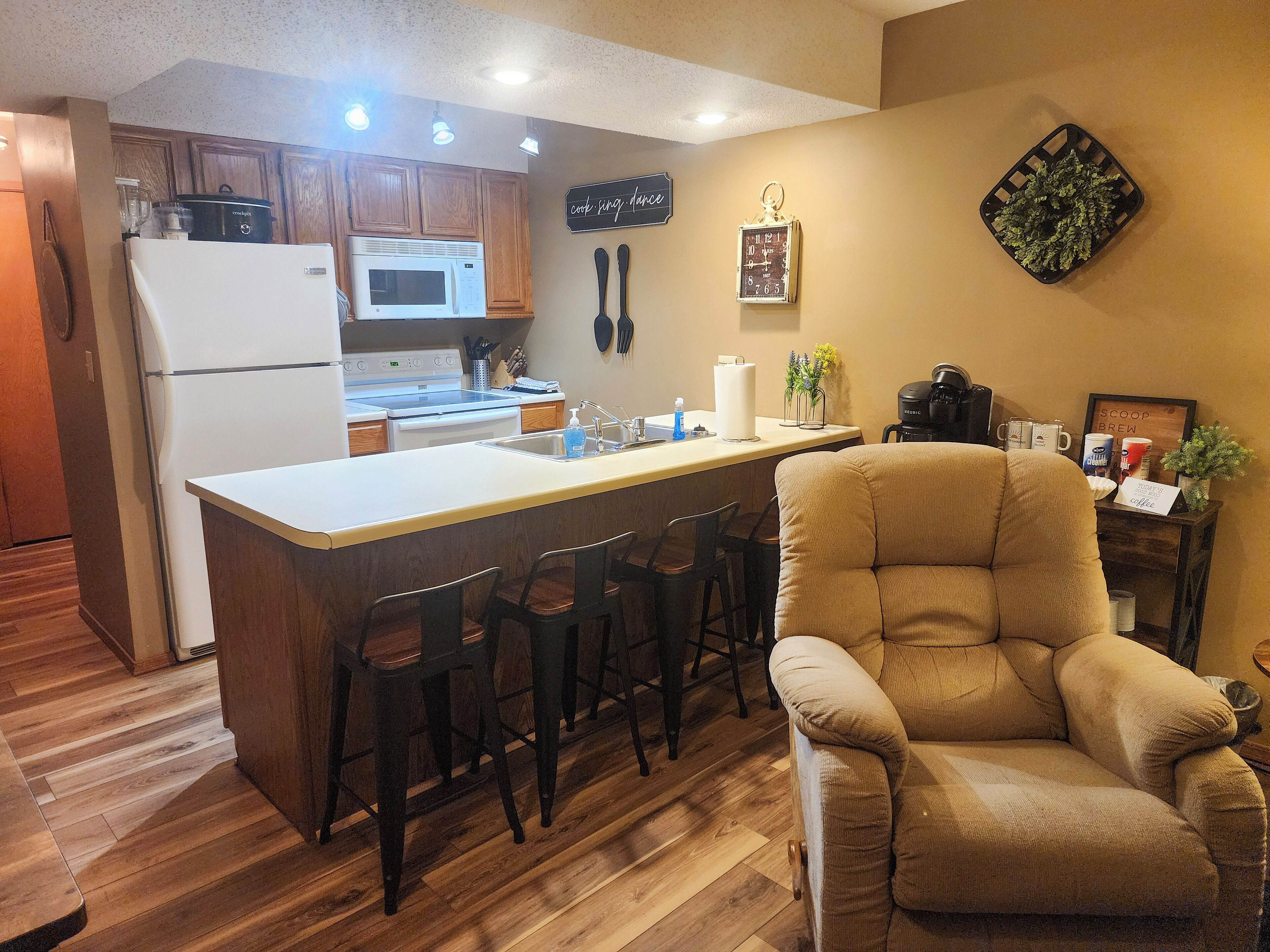 A kitchen with a central island, wooden cabinets, a refrigerator, a microwave, and a coffee machine. There are bar stools and a beige armchair.