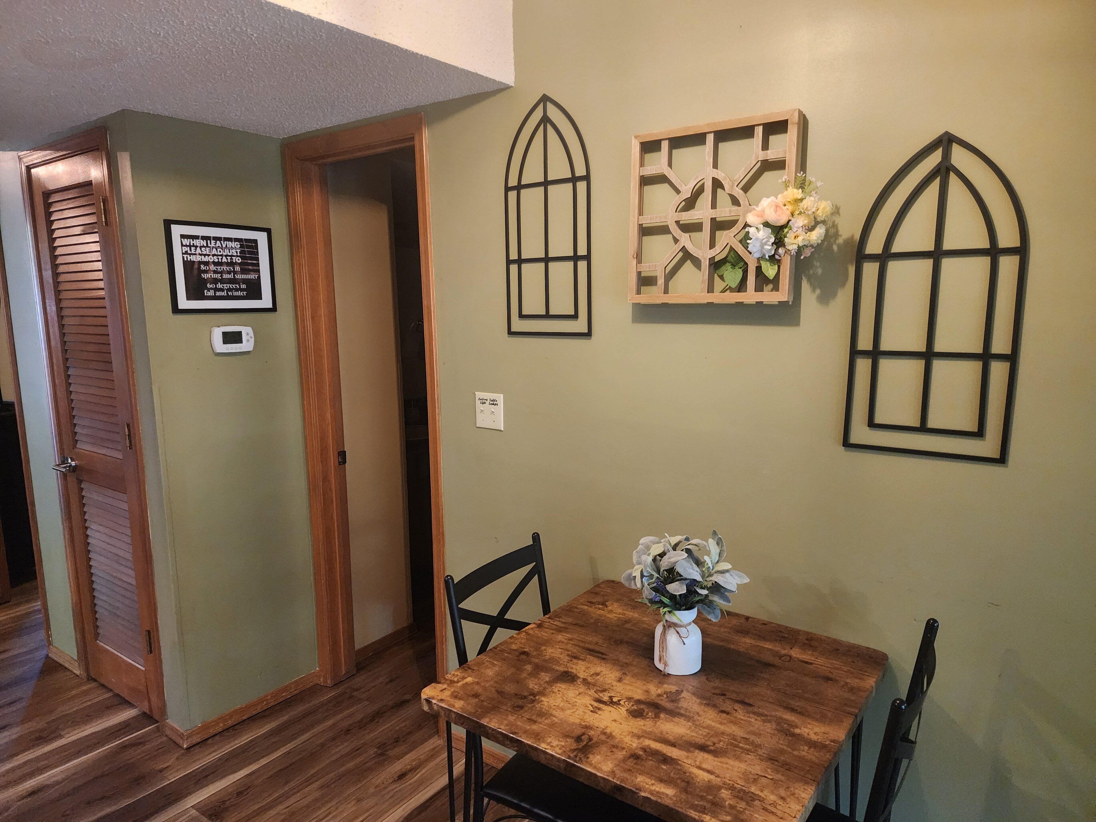 Uma sala de jantar com mesa e cadeiras de madeira, um vaso com flores e um painel decorativo que imita uma janela.